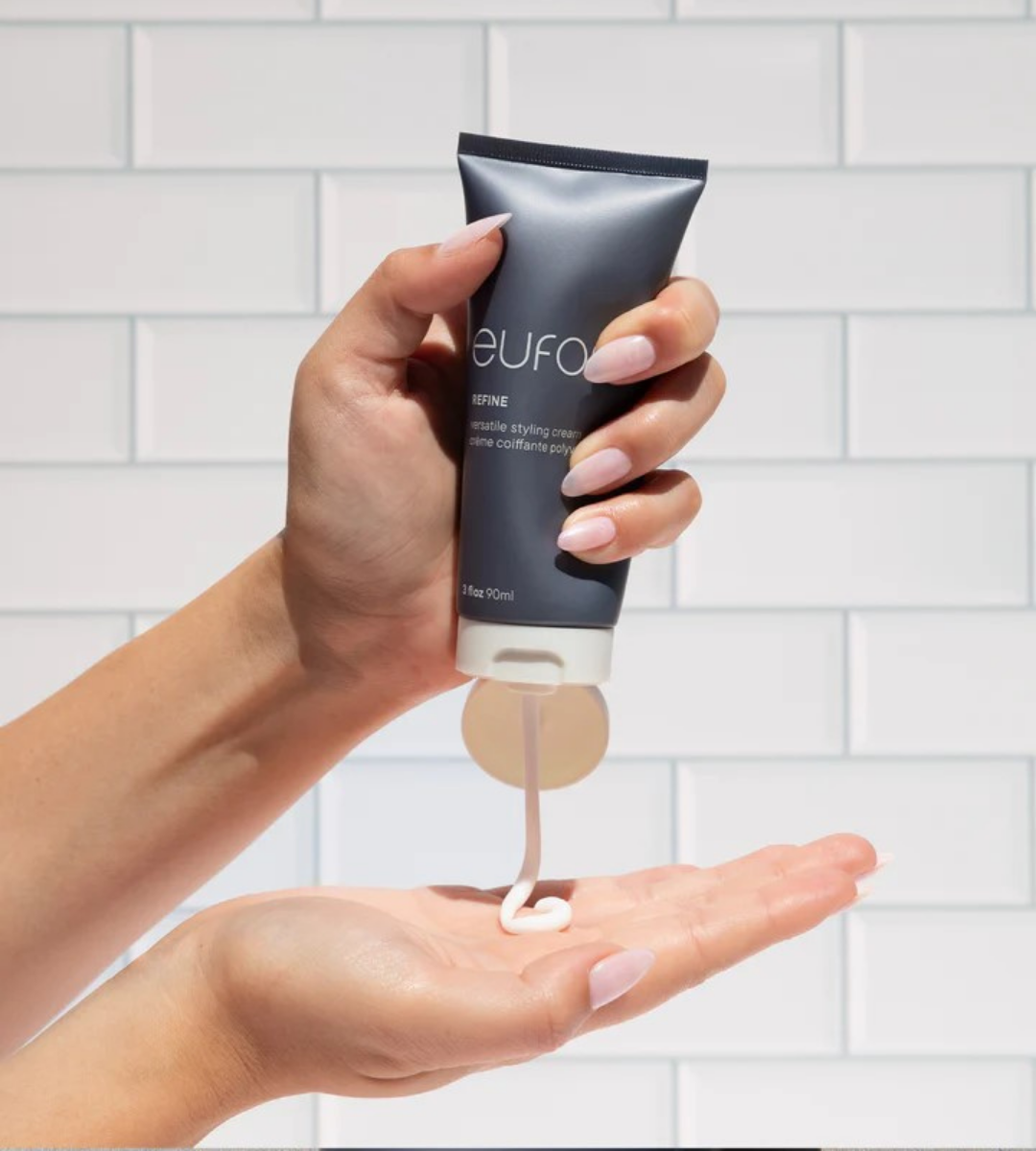 A hand squeezing Eufora Refine versatile styling cream from a gray tube into an open palm, with a white tiled wall in the background.