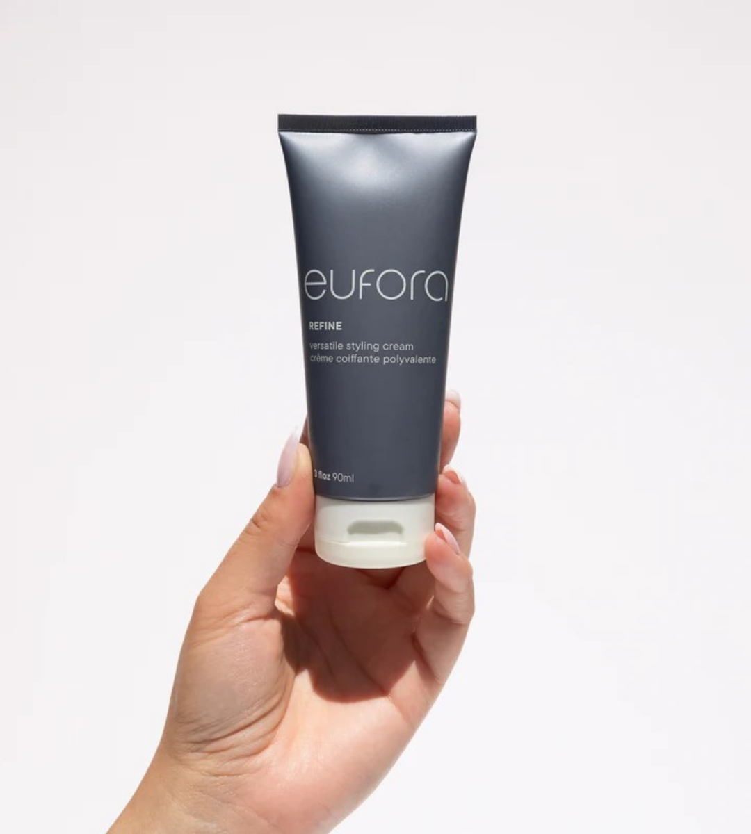 A hand holding a gray tube of Eufora Refine versatile styling cream against a plain white background, with the product label clearly visible.